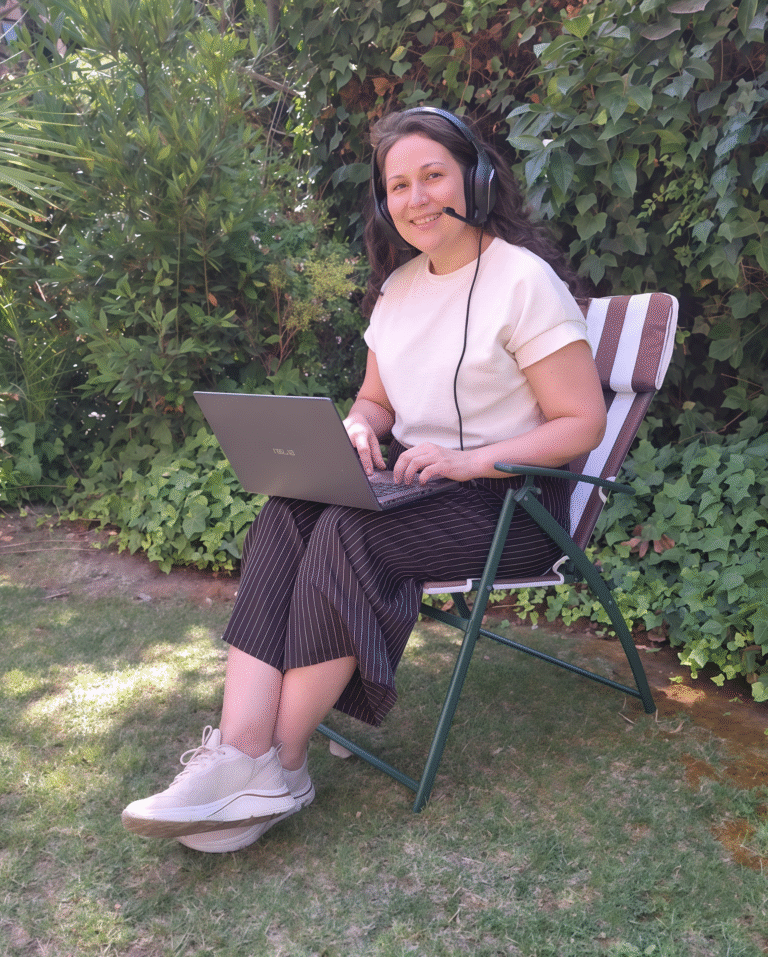 Mónica, profesora de español, sonríe mientras imparte una clase online desde su computador portátil, sentada al aire libre con audífonos y micrófono.